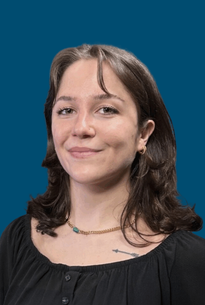 A woman with shoulder-length brown hair, wearing a black top and gold necklace, poses in front of a solid blue background.